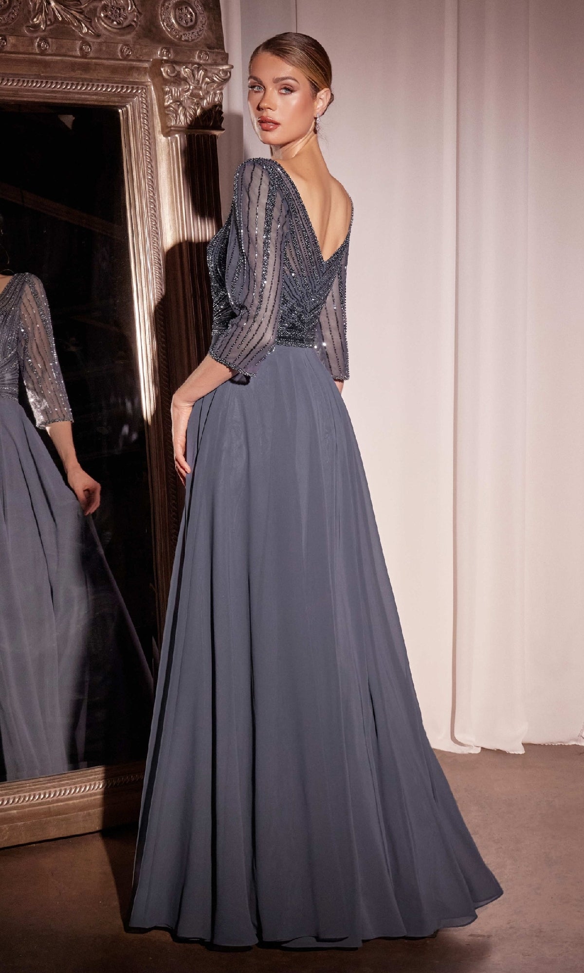 A woman in the Modest Long A-Line Formal Dress: Ladivine CC446, featuring a slate blue, floor-length design with a beaded sheer bodice and three-quarter sleeves, stands before a large ornate mirror.